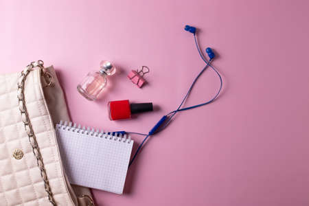 female beige clutch bag with different cosmetics and parfume containers and nail polishes on pink...
