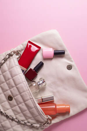 female beige clutch bag with different cosmetics and parfume containers and nail polishes on pink backgroundの写真素材