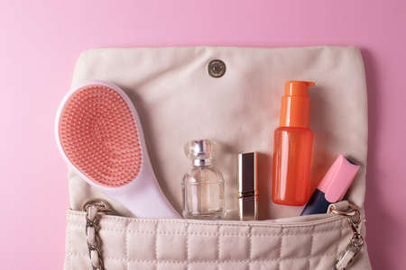 female beige clutch bag with different cosmetics and parfume containers and nail polishes on pink backgroundの写真素材