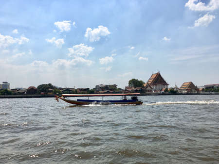 A long tail boat sailed on the Chao Phraya river and scenery of temple and house river side in Bangkok, Thailandのeditorial素材
