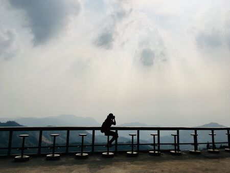 A woman taking photo on a chair and table terrace with mountain view, Sky sunlight silhouette sceneの写真素材