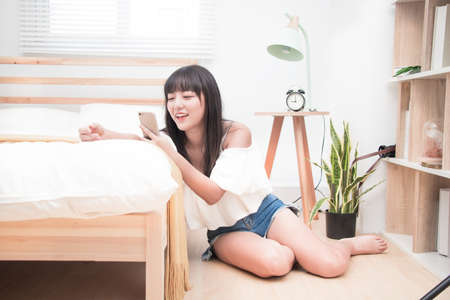 Young happy asian woman with mobile phone sitting on floor near bed at home.の写真素材