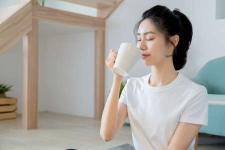 Young asia woman with ponytail in white t-shirt holding a cup of coffee or tea relaxing at home in...