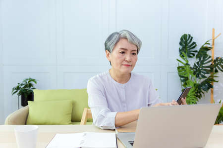 Senior Asia woman is working from home, using laptop and smart phone.
