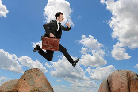 Businessman with briefcase jumping from rock to rock with clouds in backgroundの写真素材