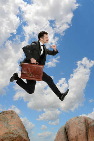 Businessman with briefcase jumping from rock to rock with clouds in backgroundの写真素材