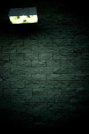 A wall lamp casts light on the rough stones of a dark wall.の写真素材