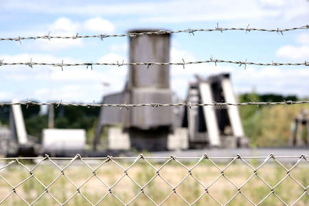 The close-up of a fence with barbed wire in front of a factory site with large factory plants.