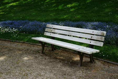 A park bench stands on the edge of a garden bed on a gravel path in the park.                       ...