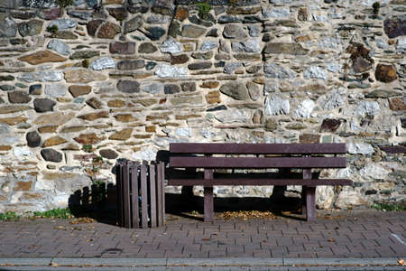 A plastic park bench and a garbage bin stand in front of a striking and rugged wall of fieldstone.
