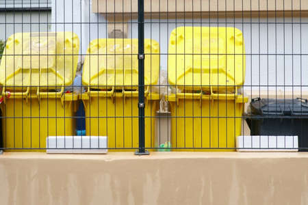 Yellow garbage cans stand in a row behind a mesh fence.の写真素材