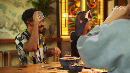 Company of multicultural friends meet in a cafe, they play in word game with phones