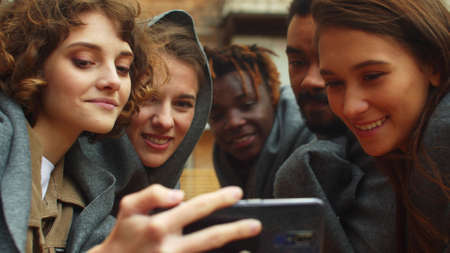 Slow motion, friends in blankets sit outside the cafe and look at photo on phoneの写真素材