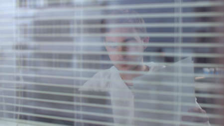 View through the blinds, man in protective workwear sit and work with computer and documents in the laboratoryの写真素材