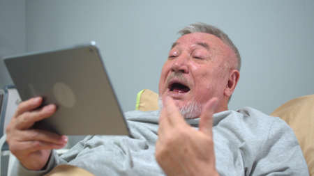 Man lie on the bed in the hospital and talk with someone on tablet emotinally
