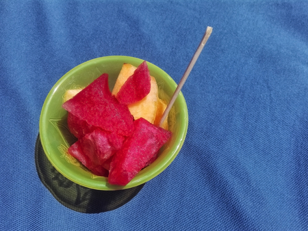 fresh watermelon and peach slices in a yellow plastic cup on a blue surfaceの写真素材