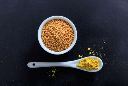 Mustard seeds and powder in porcelain bowl and spoon, black backgroundの写真素材