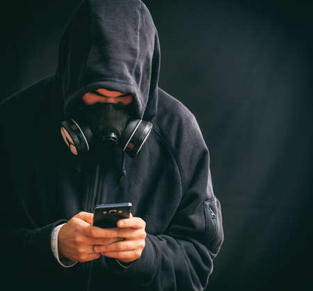 Hooded man with respiratory mask holding a smartphone on black backgroundの写真素材