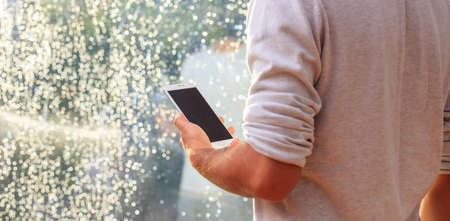 Young man infront of a window, holding a smartphoneの写真素材