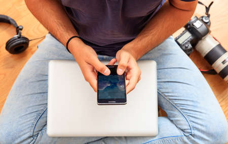 Modern workspace at home. Young photographer holding a smartphone, sitting on wooden floorの写真素材