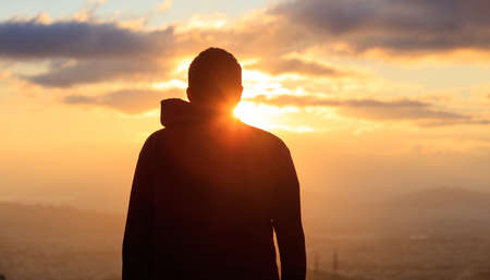 Silhouette of a young man standing alone on the top of a mountain admiring the golden sun and the city.の写真素材