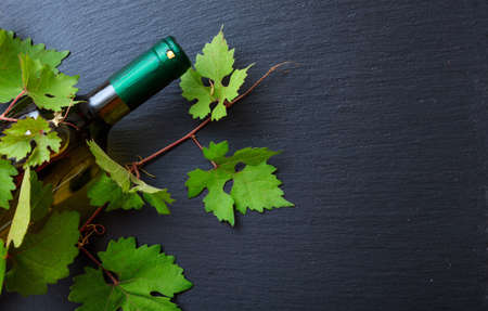 White wine bottle and fresh grape leaves on black background, top view, copy space