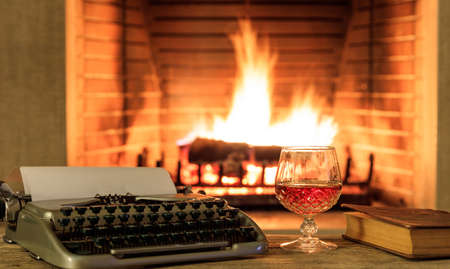 Glass of cognac and a typewriter on a wooden table, blur burning fireplace backgroundの写真素材