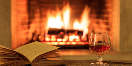 Glass of cognac and a book on a wooden table, blur burning fireplace backgroundの写真素材