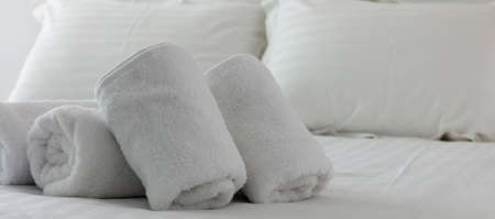 Hotel's bedroom. White fluffy, rolled towels, linen sheets and pillows on a tidy bed. Close up view.の写真素材