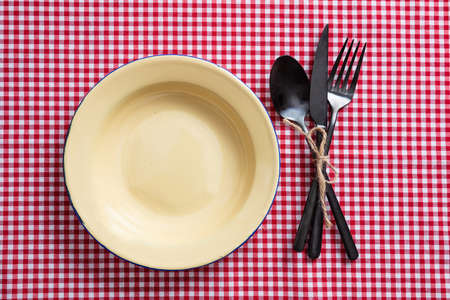 Rustic place setting. Empty enamel plate and cutlery on red checkered tablecloth, top viewの写真素材