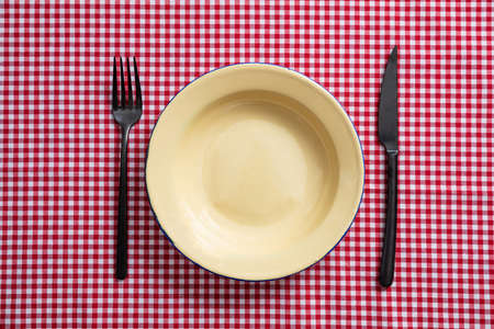 Rustic place setting. Empty enamel plate, fork and knife on red checkered tablecloth, top viewの写真素材