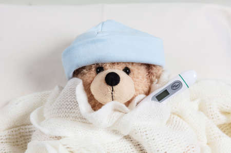 Fever, cold, flue concept. Cute teddy boy with a thermometer in bed, covered with a warm blanketの写真素材