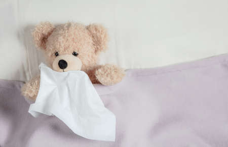 Cold, flue or allergy concept. Cute teddy in bed, covered with a warm blanket, holding a tissueの写真素材
