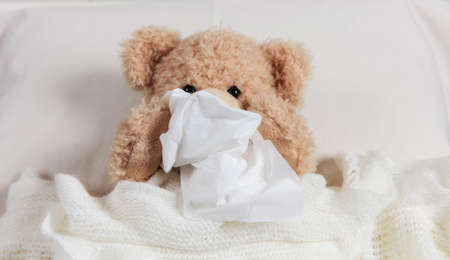 Cold, flue or allergy concept. Cute teddy in bed, covered with a warm blanket, holding a tissueの写真素材