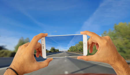 Driving and phones concept. Hands holding a smartphone, taking a photo on blur asphalt road background. 3d illustrationの写真素材