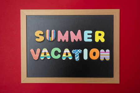 Summer vacation concept. Black chalkboard with wooden frame, text summer vacation in colorful letters, red wall backgroundの写真素材