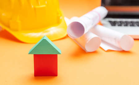 Real estate concept. Small house model, hard hat and project blueprints on orange color background