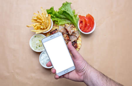 Gyros and smartphone. Hand holding a mobile phone with blank white screen, gyro pita ingredients background, copy space. Online order conceptの写真素材