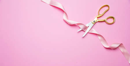 Grand opening concept. Gold scissors cutting pink satin ribbon against pink background, copy space, top view.の写真素材