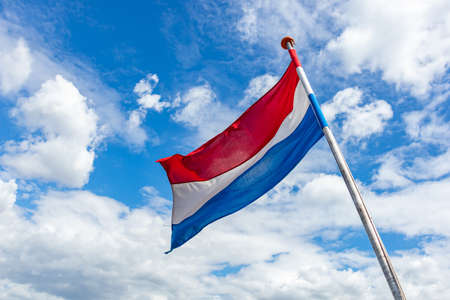 Netherlands flag. Dutch national symbol waving on pole against blue sky with clouds, sunny dayの写真素材