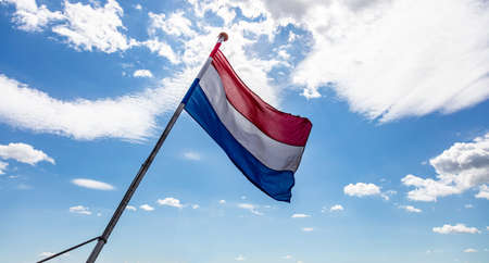 Netherlands flag. Dutch national symbol waving on pole against blue sky with clouds, sunny day. Kings or liberation day celebration conceptの写真素材