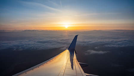 Sunset aerial view out of an airplane window. Sun falling, plane flying over the clouds. Orange color sunbeams and reflections on the plane wingの写真素材