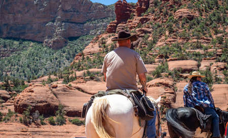 Sedona Arizona USA. May 25, 2019. People with cowboy hats riding horses back view on terrain. Red orange desert  landscape, sunny spring dayのeditorial素材
