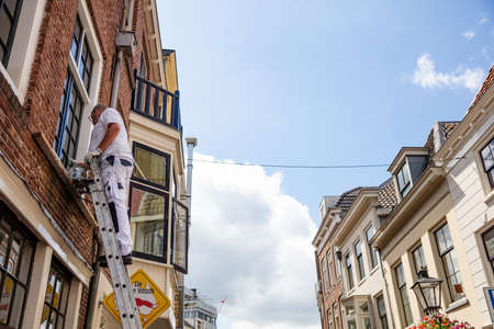 Utrecht, Netherlands - JULY 01, 2019: From below side view of male worker in white uniform standing on ladder cleaning window of old buildingのeditorial素材