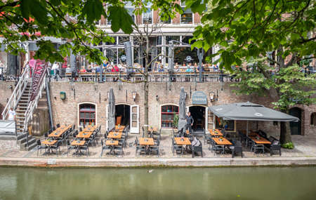 Utrecht, Netherlands - JULY 01, 2019: Waiter standing in street cafe with sunshade by river and people relaxing on benchのeditorial素材