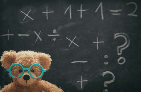 Maths concept. Smart kid in school class, cute teddy wearing blue eyeglasses and math symbols on blackboardの写真素材