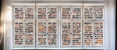 No view, loneliness, brick wall fence background through old window concept. Vintage white window pane with frame, handles and sill. Sunbeams on curtains, transparent glasses, banner.の写真素材