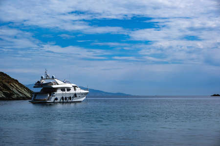 Kea, Otzias, Greece. Luxury white color yacht anchored near the land of Tzia island in the middle of the blue calm sea. Reflection of the ship, cloudy sky background.の写真素材