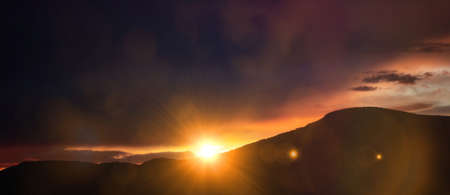 Sunset, sunrise over mountains. Sunbeams color the sky and the hills black silhouette. Sun and colorful sky background, card, wallpaper, panoramic view, banner.の写真素材