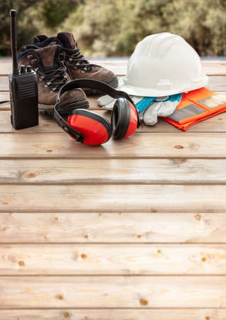 Work safety protection equipment. Industrial protective gear on wooden table, blur construction site background. Hardhat, boots, earmuffs, gloves, gogglesの写真素材